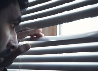 Un hombre vigila la llegada del repartidor de comida asomándose discretamente entre las cortinas como un policía escondido en un piso franco controlando quién se acerca a su perímetro
